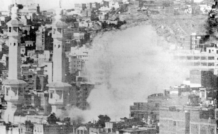 A black and white photograph showing smoke rising above the minarets of a mosque with other buildings in the background.