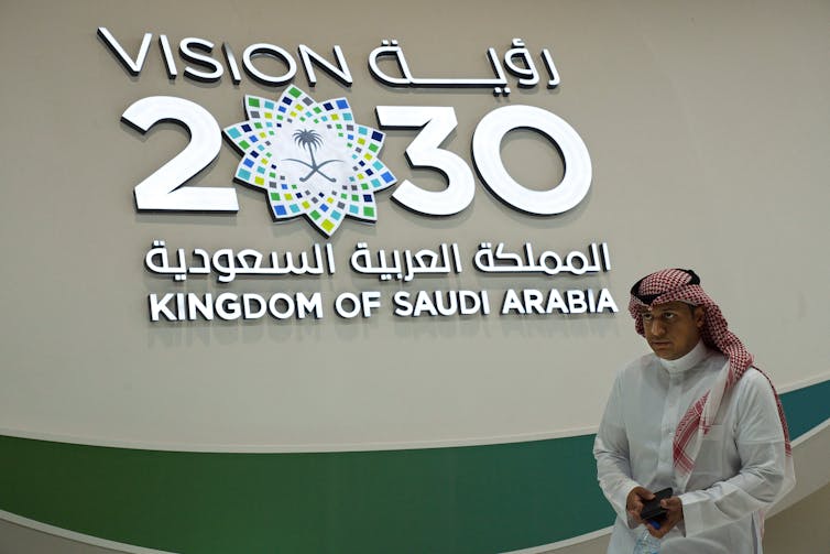 A man, wearing a headdress, walking past a display sign of 'Vision 2030.'