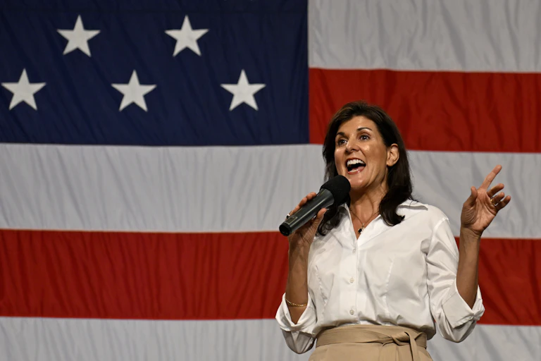 Woman gestures while speaking into microphone in front of massive American flag.