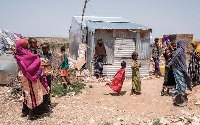 Um grupo de mulheres e crianças é fotografado em frente e ao lado de uma casa improvisada que é uma estrutura de zinco em ruínas