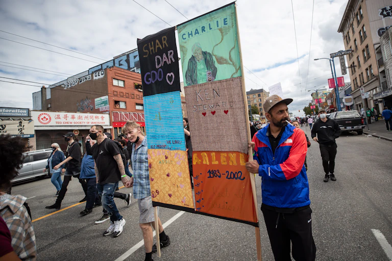 Two men marching with others, holding a banner of six squares, each with a name on it