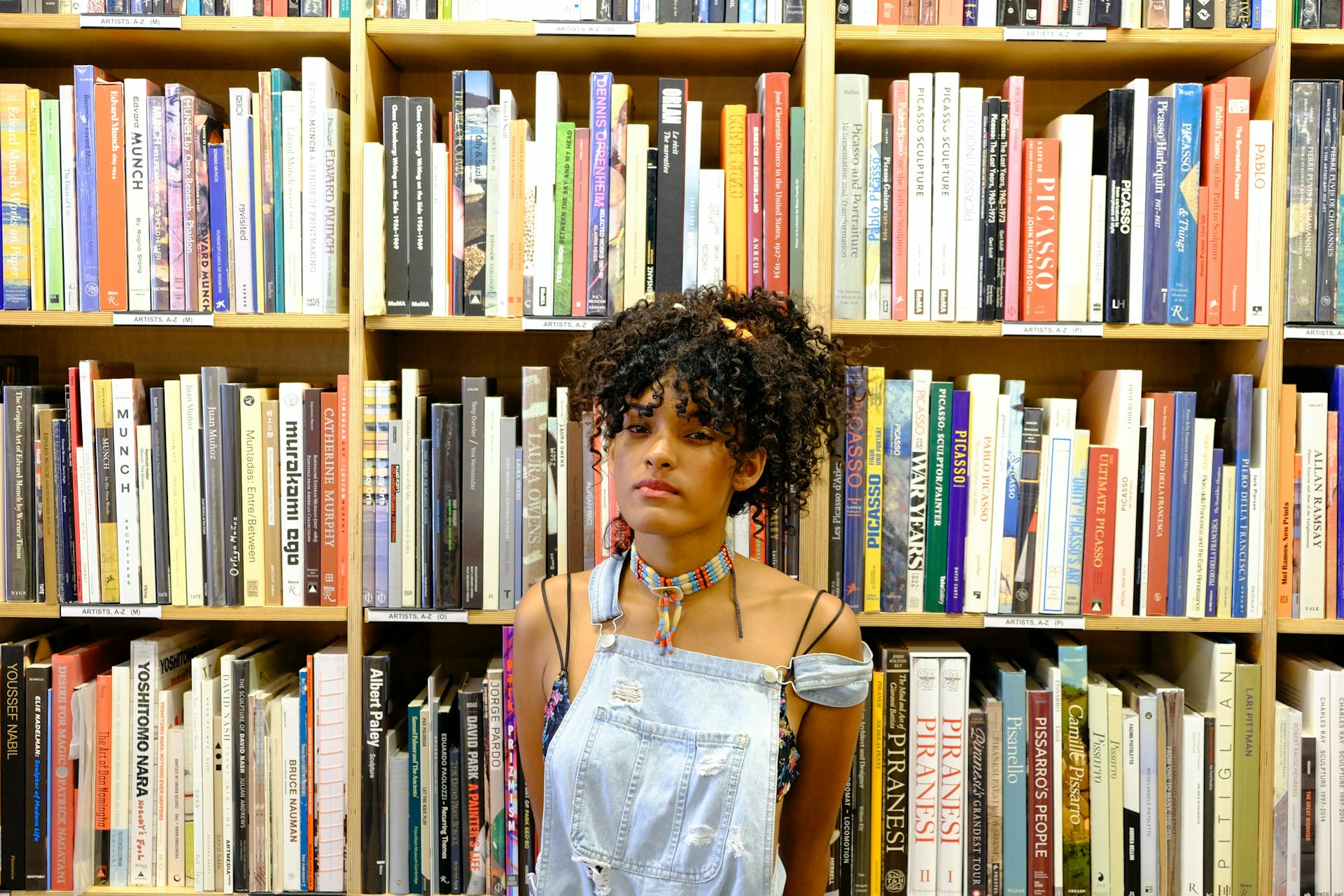 A youth in front of a bookshelf wearing overalls.