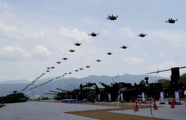 A photo showing a two long lines of quadcopter drones hovering the air above armoured vehicles.