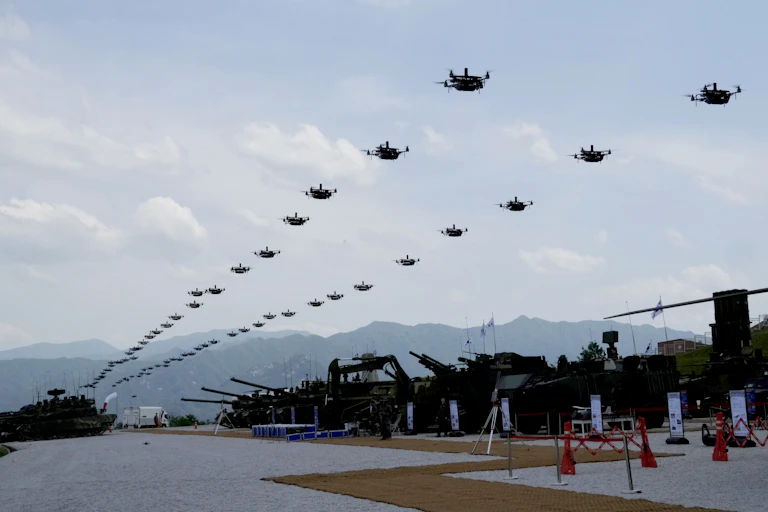 A photo showing a two long lines of quadcopter drones hovering the air above armoured vehicles.