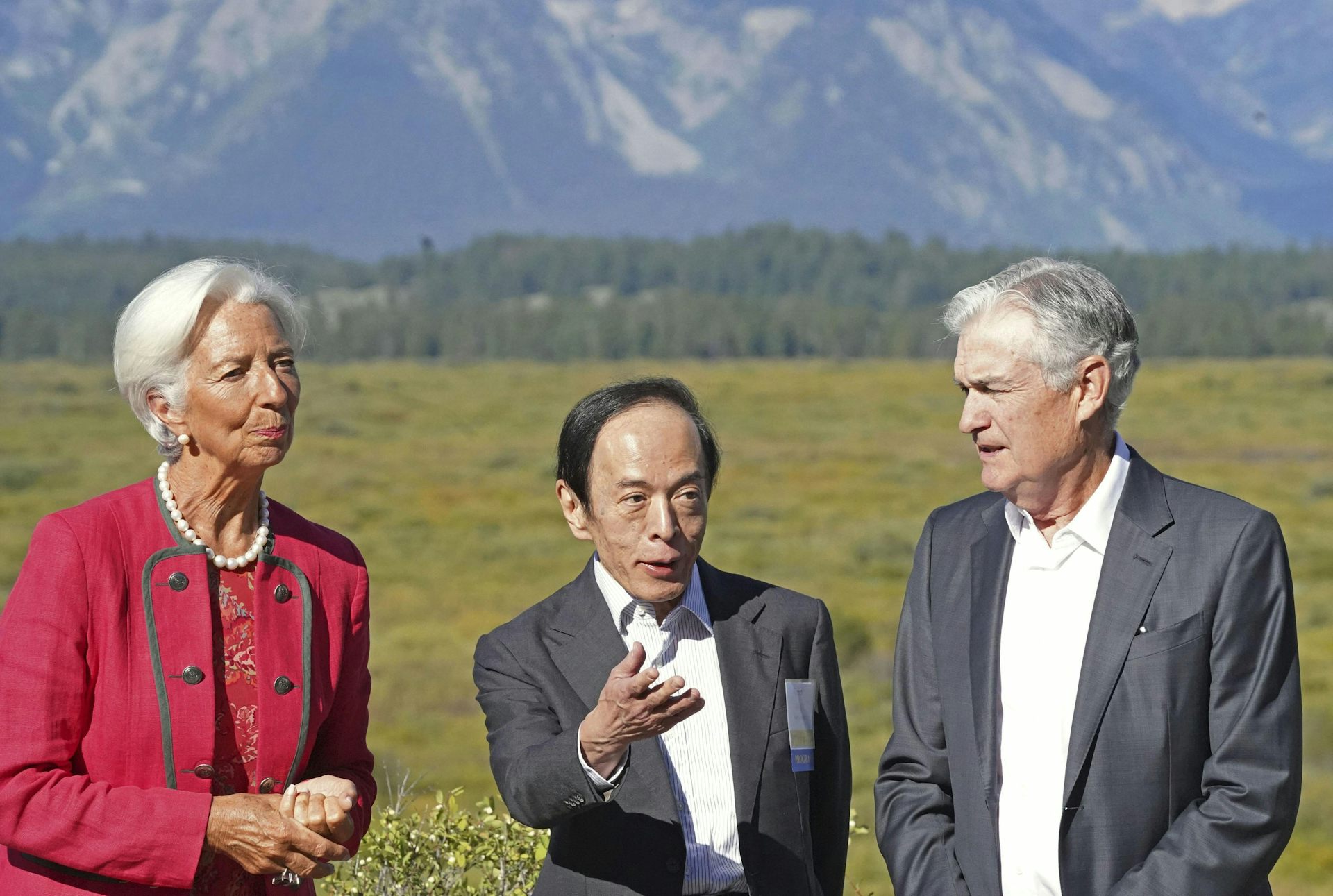 Woman and two men in suits stand in a line talking in front on green grass, trees and large mountains in the background.