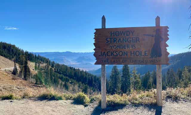 A wooden sign that says: "Howdy stranger, yonder is Jackson Hole, the last of the Old West" in front of trees and mountains, blue sky.