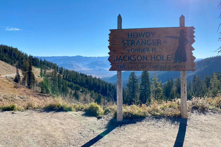 A wooden sign that says: "Howdy stranger, yonder is Jackson Hole, the last of the Old West" in front of trees and mountains, blue sky.