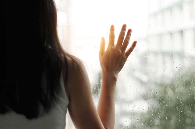 A woman stands looking out a window with her hand to rain-splattered glass.