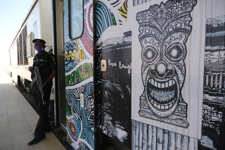 A policeman with a gun rests against a train carriage which is adorned with a graphic illustration of a traditional sculpture.