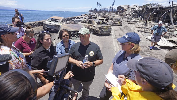 A man wearing a T-shirt with the state seal of Hawaii speaks with reporters, standing next to a woman with 'FEMA' on her cap and shirt.