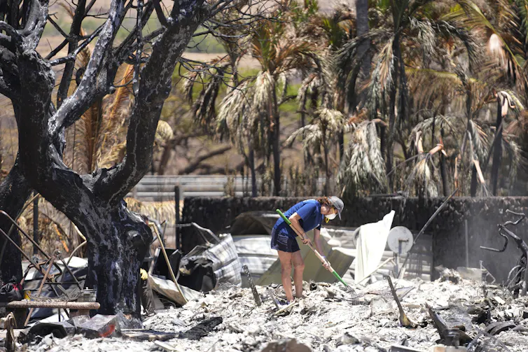 A woman wearing shorts, a T-shirt and face mask uses a pitch fork to dig through the ash of a home in Lahaina, Hawaii.