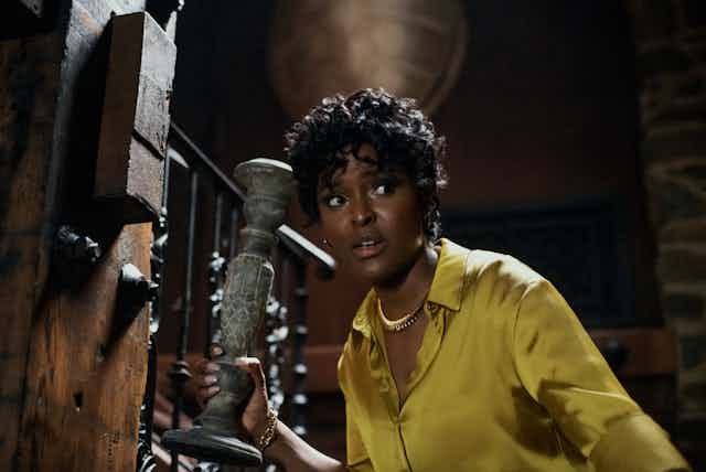 Antoinette Robertson as Lisa in The Blackening holding a candlestick and wearing a yellow silk blouse.