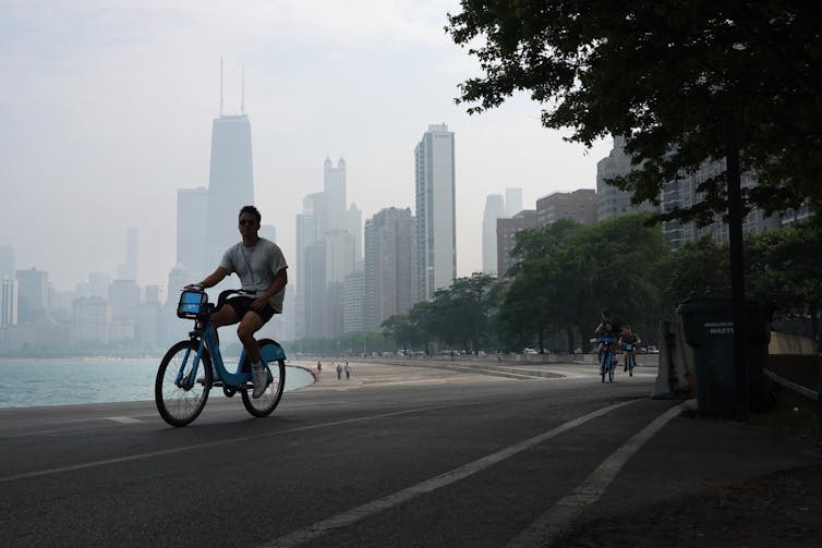 A young man rides a rental bicycle along the shores of Lake Michigan in Chicago as smoke obscures the view of the city skyline in the background.