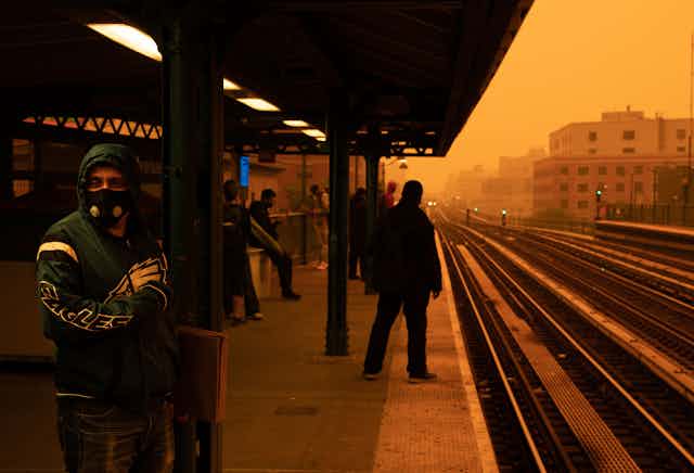 A man stands on a subway platform wearing a hooded jacket and face mask to cover as much of his body as possible against the smoke particles that have turned the sky a dark orange. The buildings along the tracks quickly disappear in the haze.
