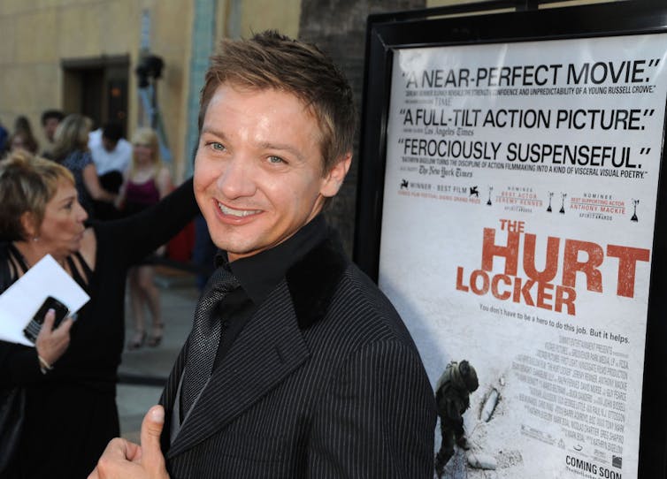 Man in black suit smiles in front of movie poster.