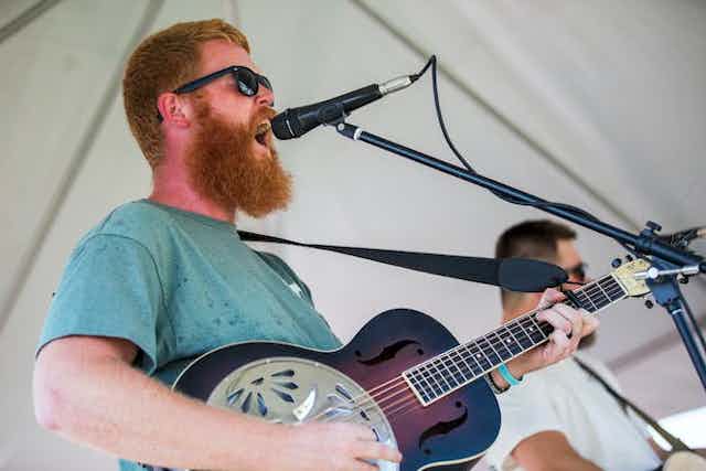 A red-headed man with a beard sings into a microphone and plays guitar.