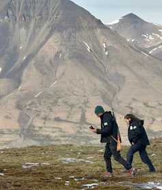 Casal caminha em área de vegetação rasteira, rodeado por montanhas