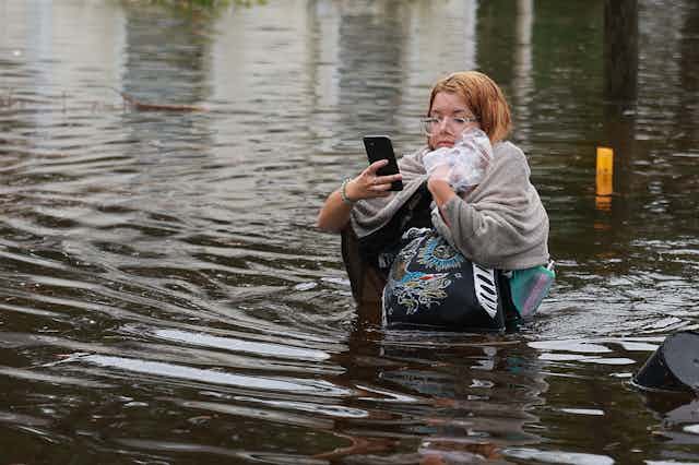 A woman holds her phone and clutches several bags, trying to keep them above the waist-high water as she wades to drier ground. A house behind her has water up nearly to its windows.