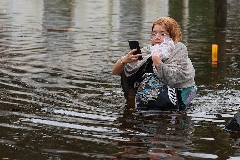 A woman holds her phone and clutches several bags, trying to keep them above the waist-high water as she wades to drier ground. A house behind her has water up nearly to its windows.
