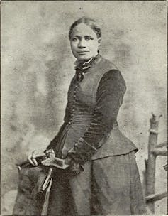 A black and white photograph of a woman in dark clothing standing formally while holding on to a chair.
