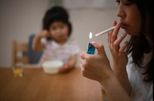 A mother lights up a cigarette near her toddler, who sits in a high chair in the background.