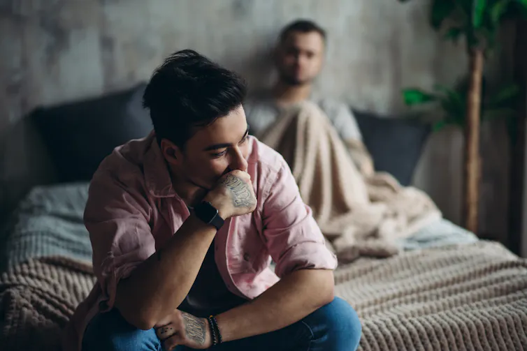 A man sits on the edge of a bed with a troubled look on his face. Another man sits on the bed behind him.