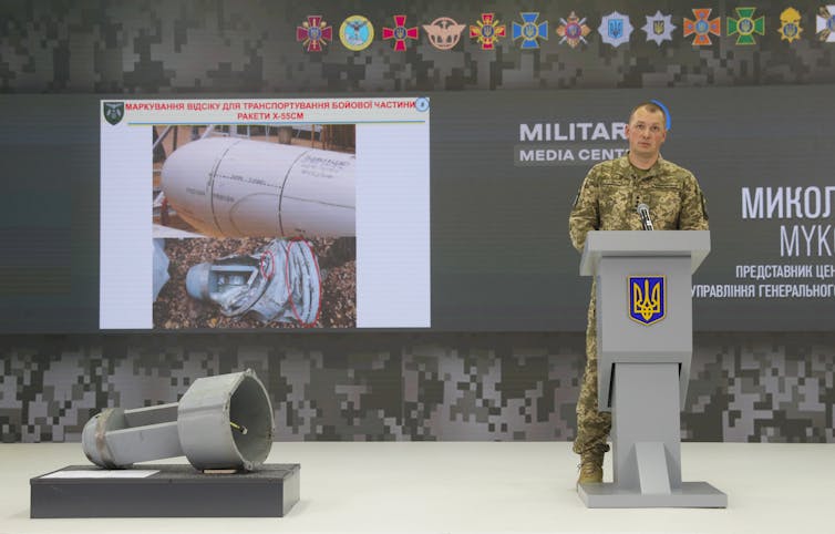 A Ukrainian soldier stands at a lectern with a piece of a Russian missile and a screen showing the fully constituted missile.