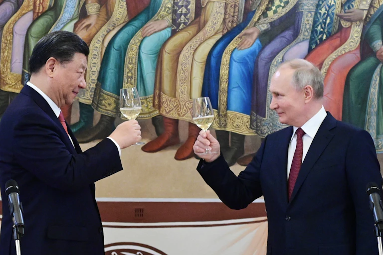 Two men in dark suits raise champagne glasses to one another. An ornate mural is behind them.