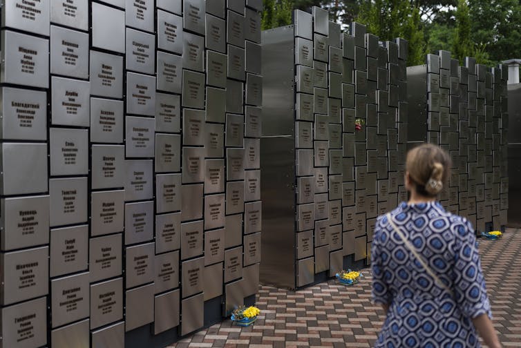 A woman in a blue patterned dress looks at a large memorial wall with Ukrainian names etched on it.