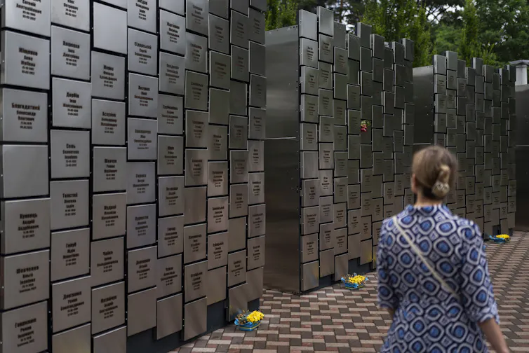 Une femme vêtue d’une robe bleue à motifs regarde un grand mur commémoratif sur lequel sont gravés des noms d’Ukrainiens