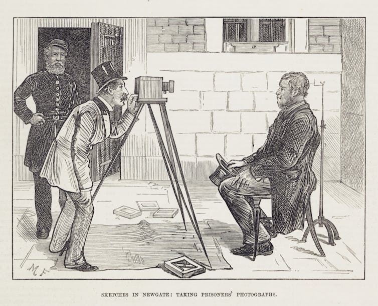 A black and white drawing shows a person taking a photograph of a seated man on an old looking camera,