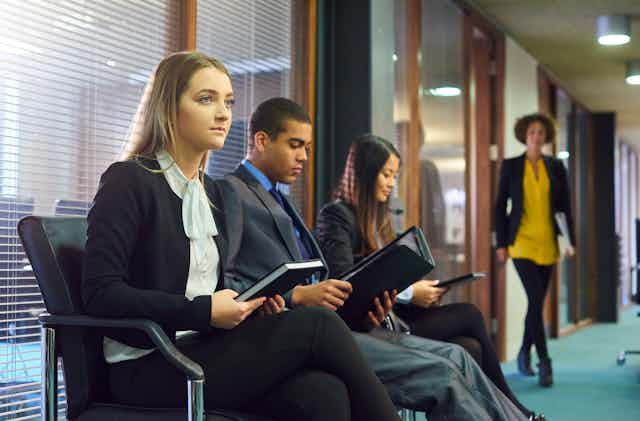 Three interview candidates in professional attire wait nervously in the reception area of an office building. A member of the interview panel can be seen walking toward them.