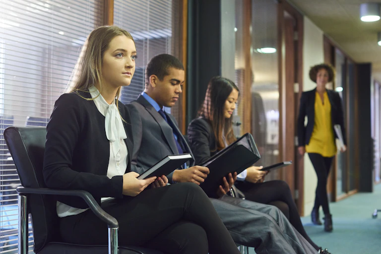Three interview candidates in professional attire wait nervously in the reception area of an office building. A member of the interview panel can be seen walking toward them.