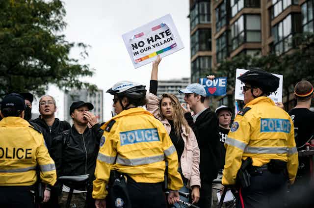 Police in yellow jackets stand in a line in front of a group of people. A woman in a pink jacket holds a placard reading: No hate in our village.
