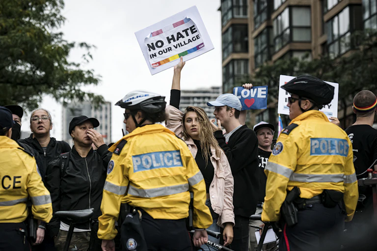 Police in yellow jackets stand in a line in front of a group of people. A woman in a pink jacket holds a placard reading: No hate in our village.