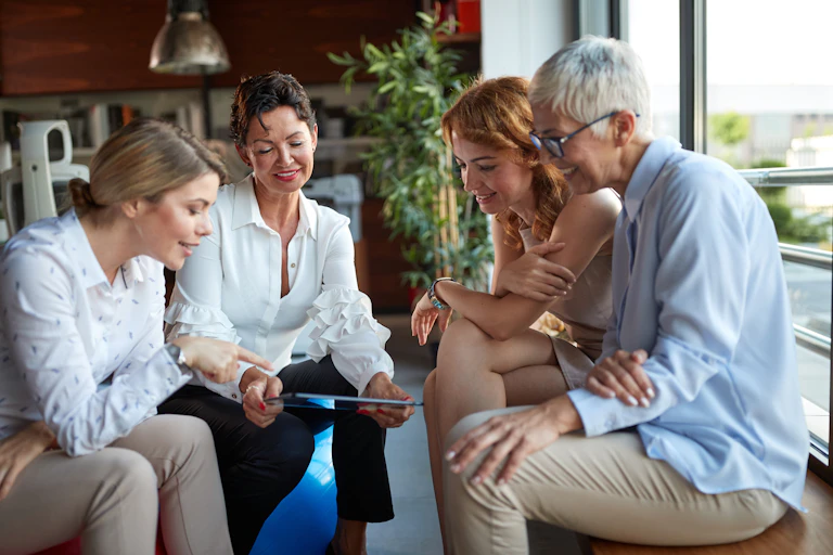 A group of women of different ages smiling and looking at a tablet, pot plants, window, shelves in the background.