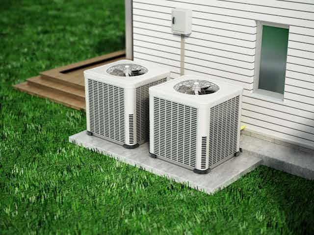 Situated on a concrete slab amidst a grassy lawn, two home heat pumps stand next to an exterior house wall.