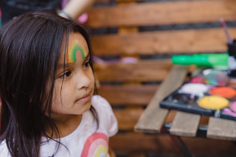 A young child stands next to a set of paints with a rainbow painted on her forehead.