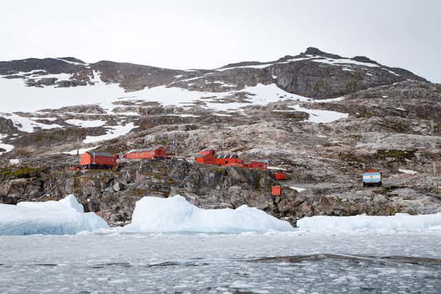 Red buildings of a research station on rocky slope down to icy sea with snow-capped mountains behind