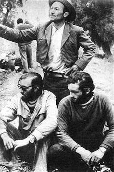 Photograph of two seated boys with a standing man talking to the cameras.