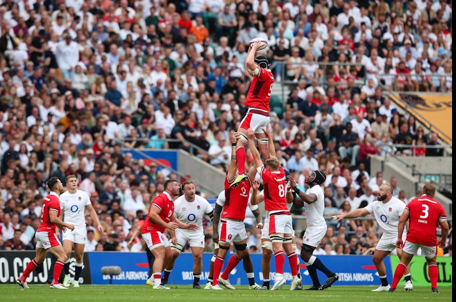 A group of rugby players in red hoist one player above a group of rugby players in white. 