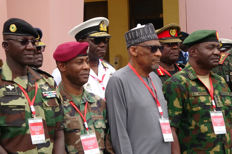 A man wearing traditional attire with a cap, standing in the midst of men in military uniform.