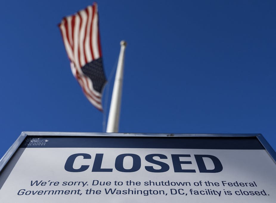 A "closed" sign is displayed on a government building during the US government shutdown in December 2018. An American flag is visible in the distance.