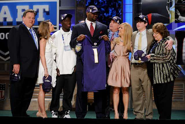 Man poses on stage wearing purple hat and holding a purple jersey surrounded by men and women.