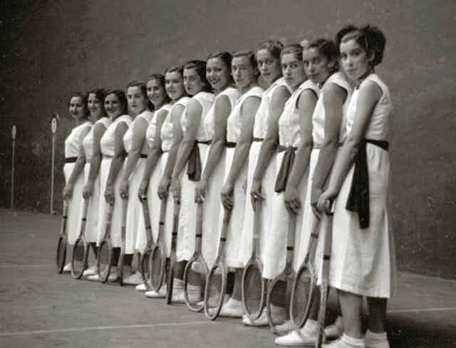Grupo de mujeres vestidas de blanco posando con las raquetas apoyadas en el suelo.