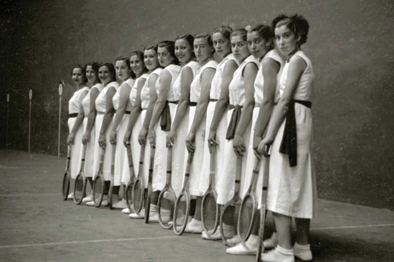 Grupo de mujeres vestidas de blanco posando con las raquetas apoyadas en el suelo.