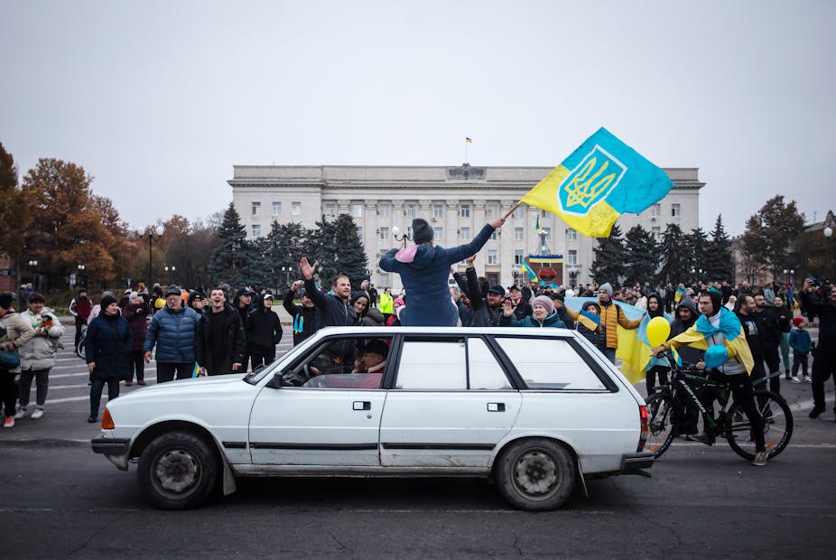 A woman, sitting on top of a white car, waves a Ukrainian flag as a large group of people cheer her on