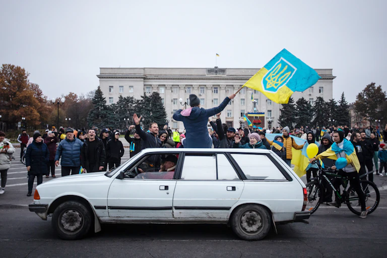 A woman, sitting on top of a white car, waves a Ukrainian flag as a large group of people cheer her on