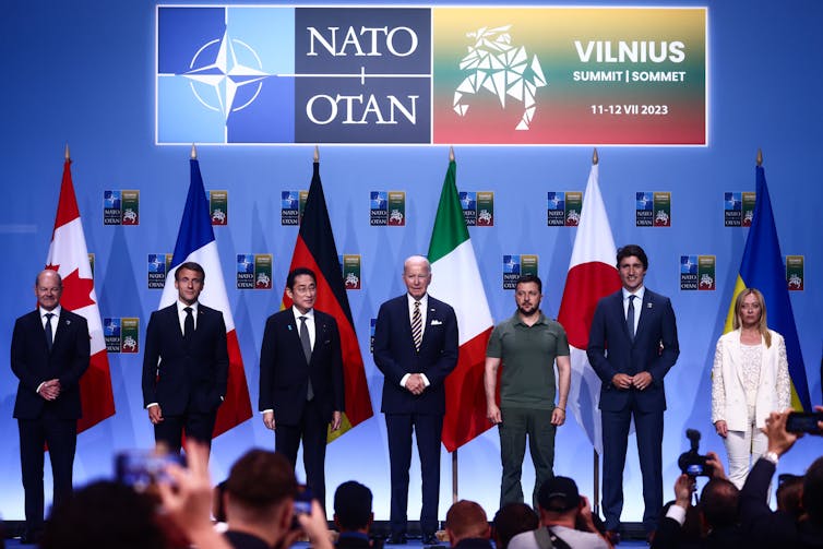 Five men in dark suits and a woman in a white suit stand with a man in shirt and pants before standing flags of various nations.