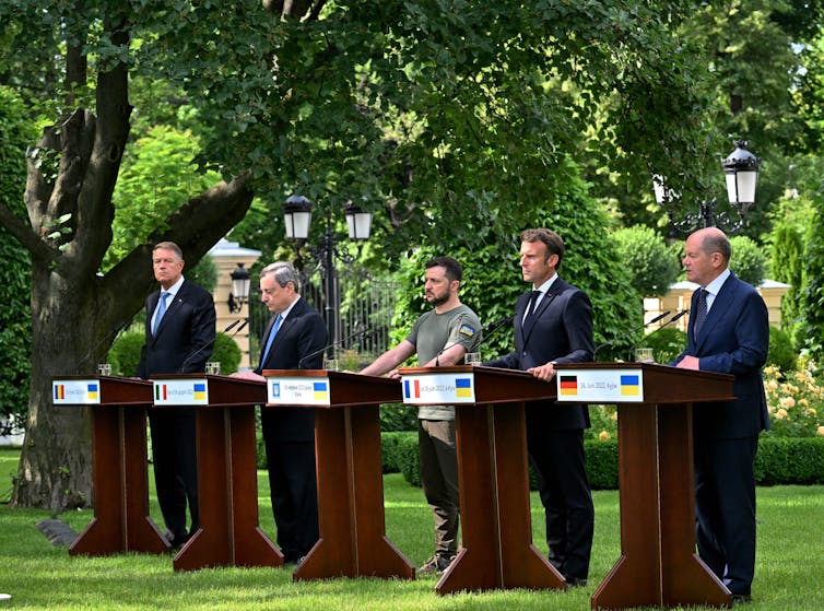 Five men stand in an outside green space, behind individual lecterns.